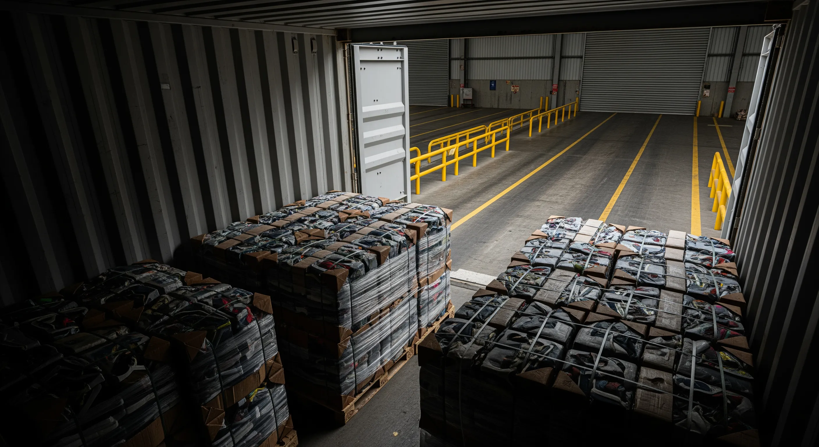 Low-angle view from inside a container looking out onto a loading bay with stacked wrapped bales and dock leveler in an export warehouse.